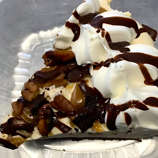 A slice of pie with whipped cream, chocolate drizzle, and nuts in a plastic container.
